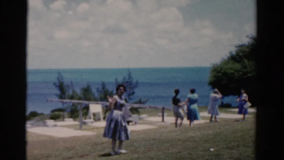 1975 Florida 8mm frame of women scattered across a grassy seaside overlook, with the ocean and trees behind them.