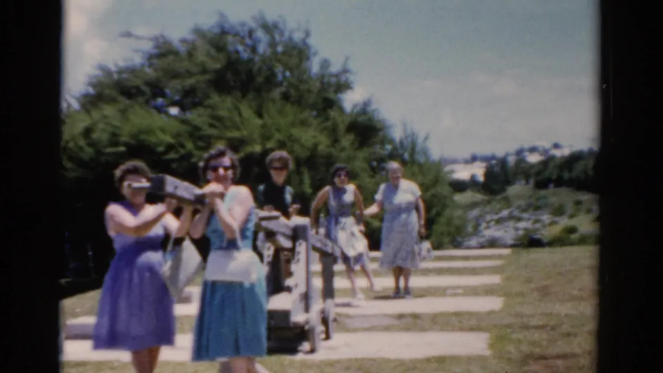 1975 Florida home-movie frame of women gathered near a seaside path, with one person holding a camera-like object toward the filmer.