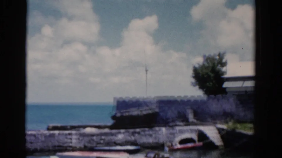 1975 Florida coastal view with blue water, a low seawall, a small boat near the edge, and clouds over the horizon.