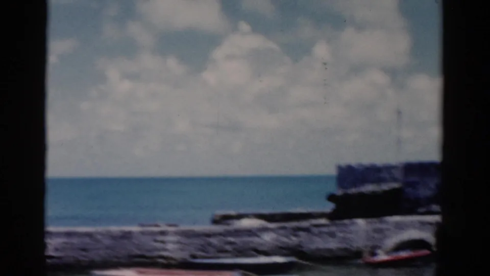 1975 Florida shoreline view with open water, clouded sky, stone edging, and a small boat partly visible near the frame edge.