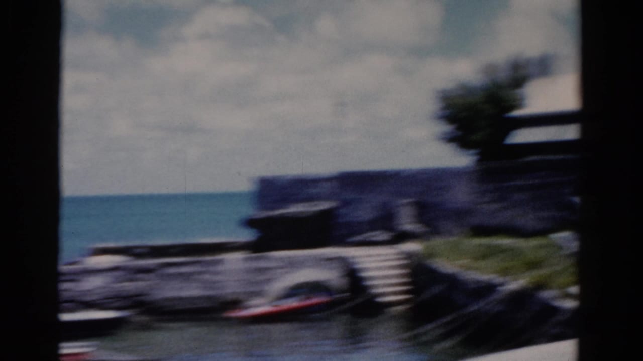Blurred moving view of blue water, small boats, a low wall, and shoreline trees.