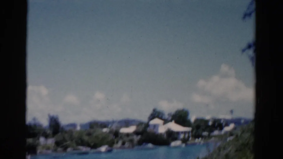 1975 Florida 8mm frame looking across blue water toward low shoreline houses, trees, and a partly cloudy sky.