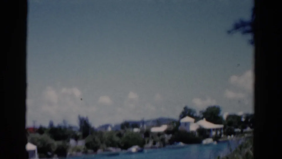 1975 Florida 8mm frame of a waterfront landscape with blue water, low houses, trees, and partly cloudy sky from a moving car.