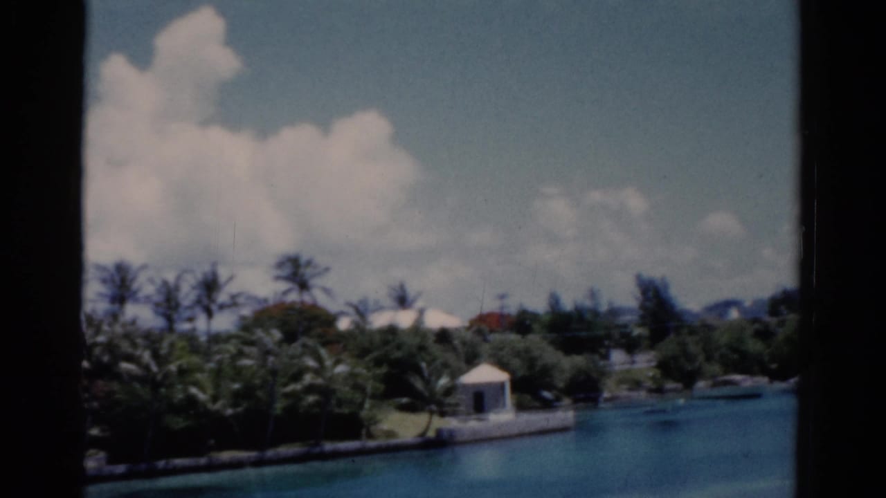 1975 Florida 8mm frame showing a palm-lined waterfront resort area with turquoise water, white buildings, clouds, and dark window edges.