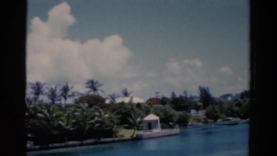 1975 Florida 8mm frame showing a palm-lined waterfront resort area with turquoise water, white buildings, clouds, and dark window edges.