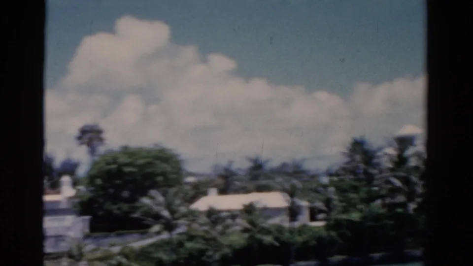 1975 Florida 8mm view of low white-roofed resort buildings behind palms and broad tropical trees under a bright clouded sky.