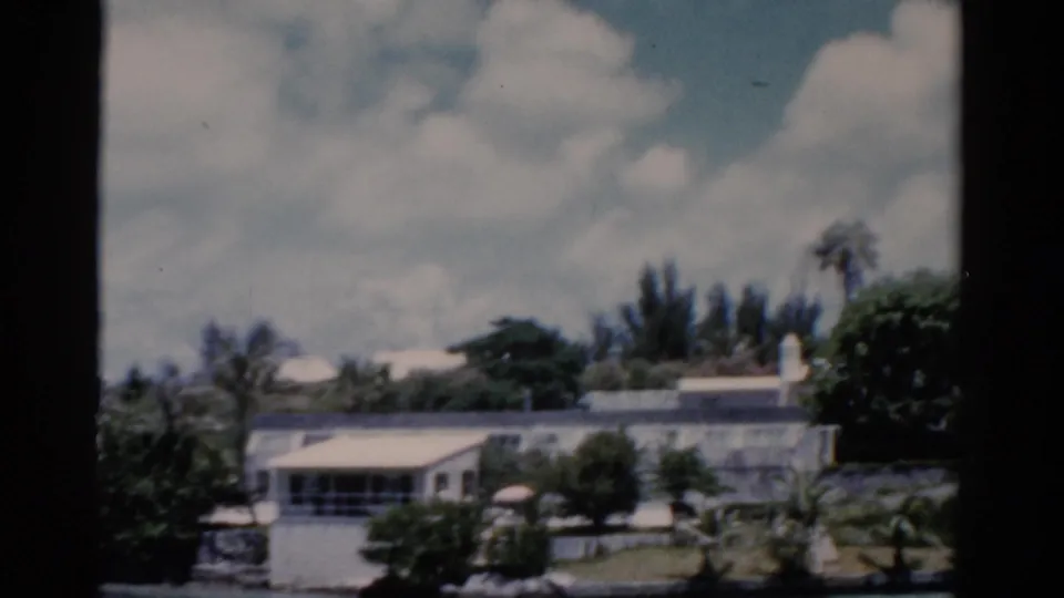 1975 Florida resort scene with a low white building, palms, trimmed greenery, lawn, pale roofs, bright clouds, and dark window framing.