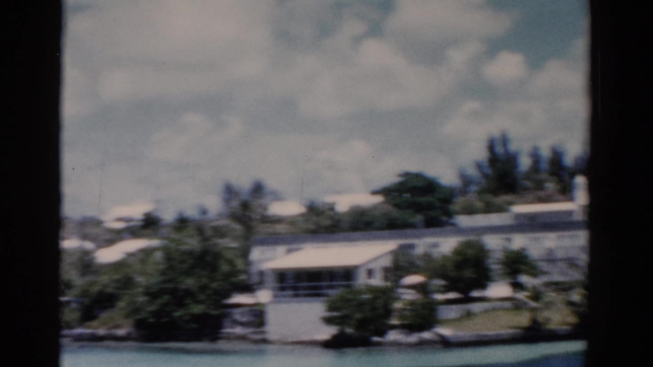 1975 Florida 8mm frame showing low white waterfront houses or mobile homes, trees, and blue water seen through film gate edges.