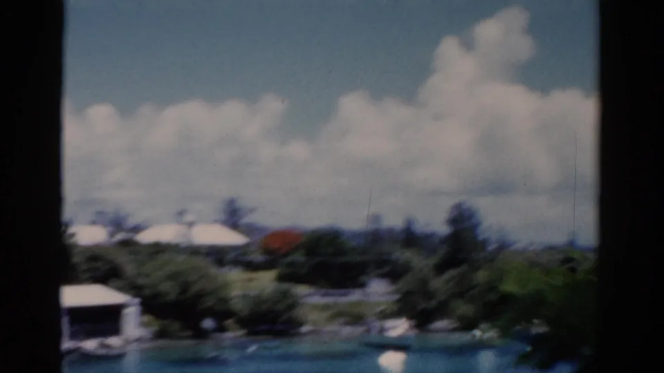 1975 Florida lakeside view with low white buildings, dense greenery, a small dock-like structure, and bright clouds over blue water.
