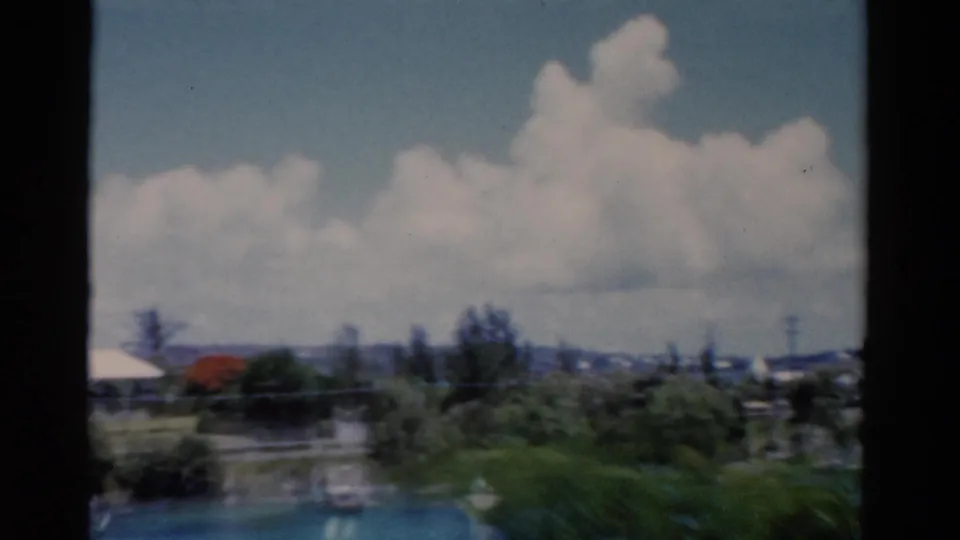 1975 Florida home-movie frame with blue water, shoreline greenery, a small boat, and distant low buildings beneath large white clouds.