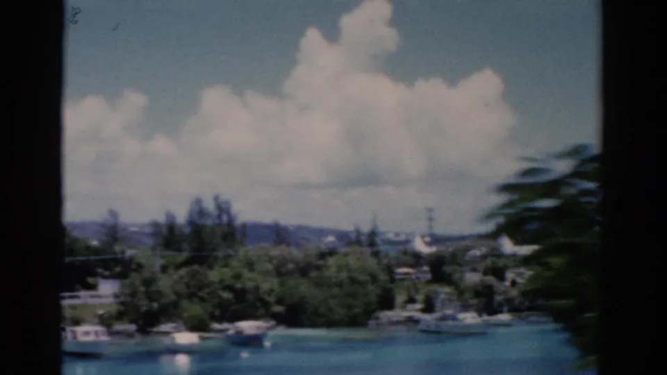 1975 Florida 8mm view of small boats on blue water, dense shoreline trees, and low residential structures under a clouded sky.