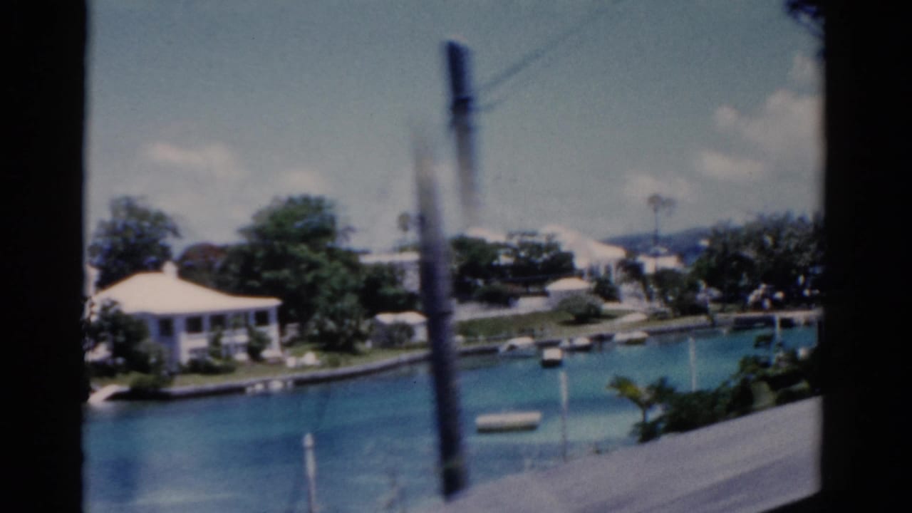 Blue bay with anchored boats, white waterfront house, trees, and a blurred foreground pole.