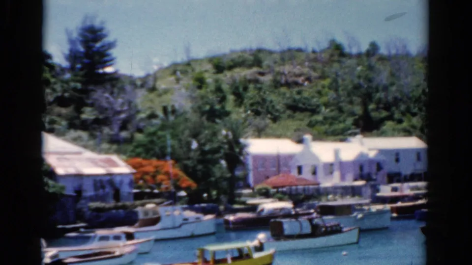 1975 Florida 8mm view of moored boats, pastel and white waterfront buildings, hillside vegetation, and a red-flowering tree.