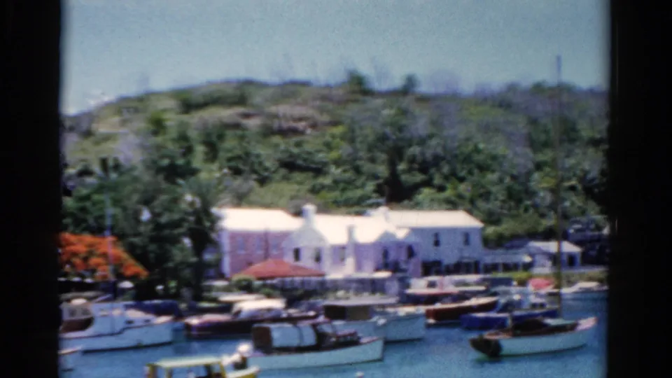 1975 Florida frame showing anchored boats in a blue bay, white waterfront buildings, docks, and lush hillside vegetation.