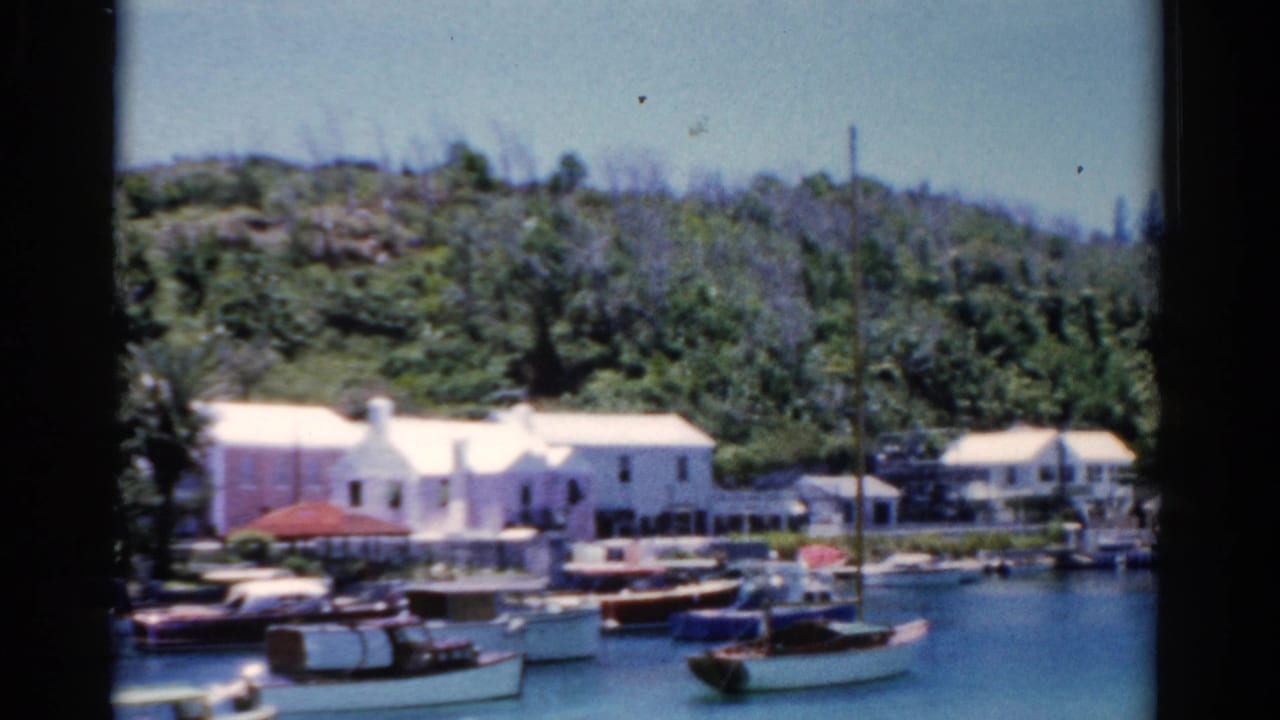 1975 Florida 8mm frame showing small boats moored near white waterfront buildings below a wooded hillside.