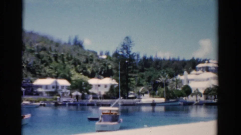 1975 Florida 8mm harbor frame showing shoreline buildings, moored boats, and tall trees along the water.