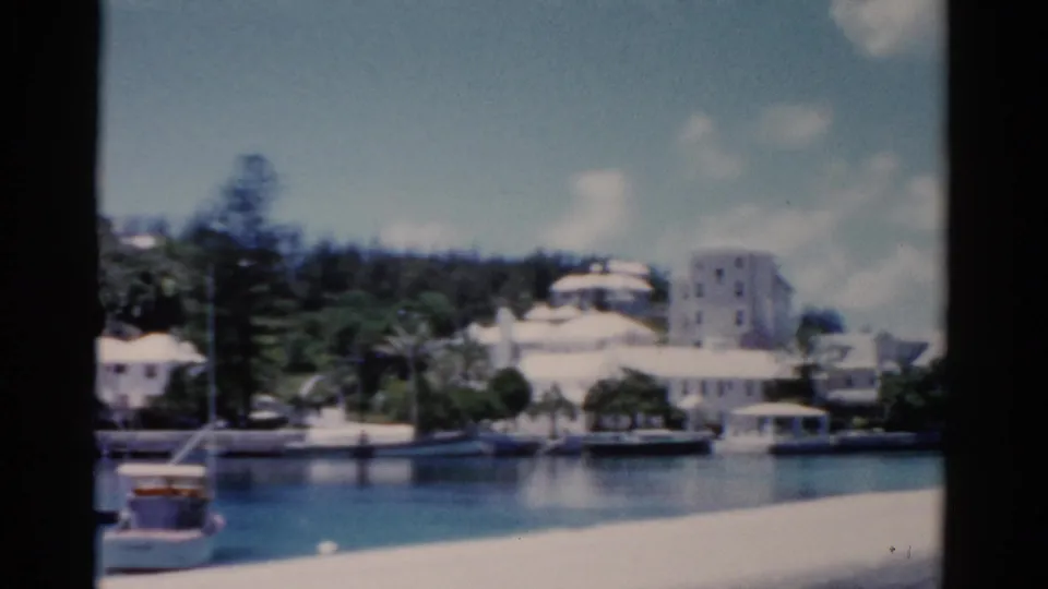 1975 Florida coastal harbor view with small boats, shoreline structures, trees, and taller white buildings in the distance.