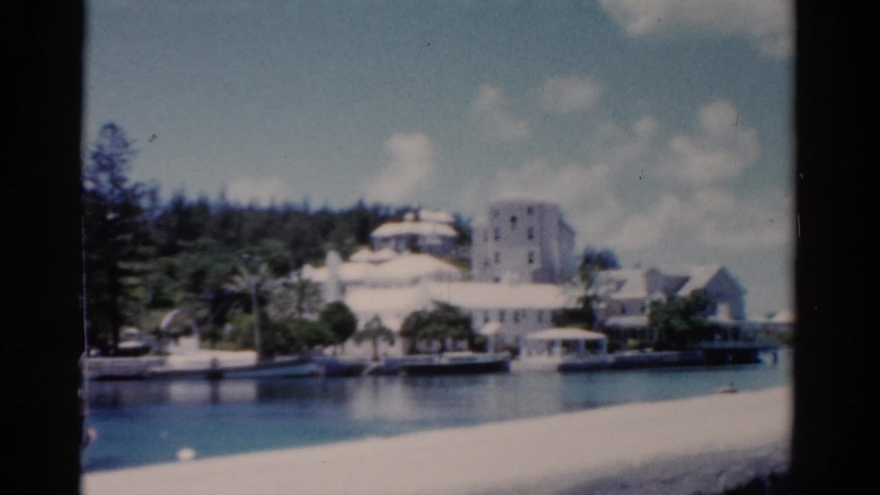 1975 Florida 8mm frame showing calm waterfront, white buildings, shoreline trees, and blue sky from a visitor viewpoint.