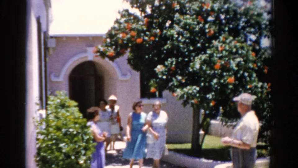 1975 Florida home-movie frame of adults gathered outside by a pale building, arched doorway, shrubs, and fruit tree.