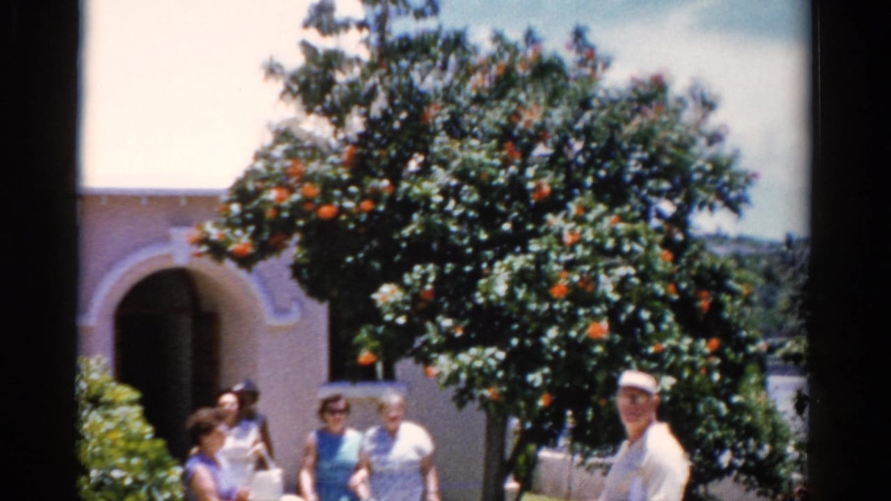 1975 Florida home-movie frame of older adults outside a white stucco church-like building, with an orange tree overhead.