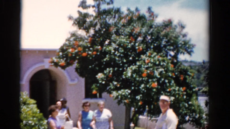 1975 Florida home-movie frame of older adults outside a white stucco church-like building, with an orange tree overhead.