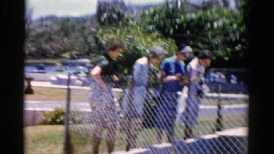 1975 Florida home-movie frame showing older adults walking past a chain-link fence after leaving the church area.