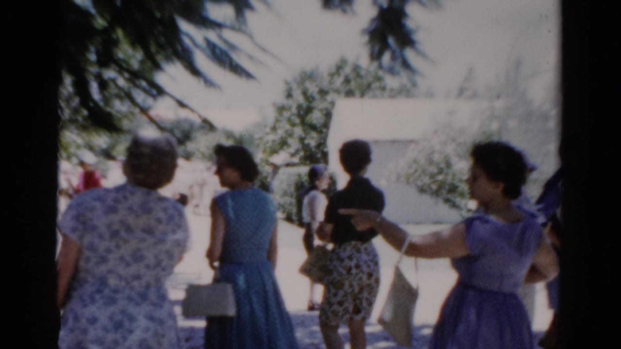 Florida 1975 home-movie frame showing adult women in patterned and blue dresses walking through a sunny park walkway.