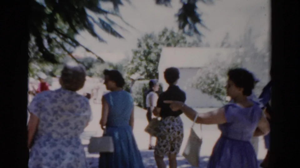 Florida 1975 home-movie frame showing adult women in patterned and blue dresses walking through a sunny park walkway.