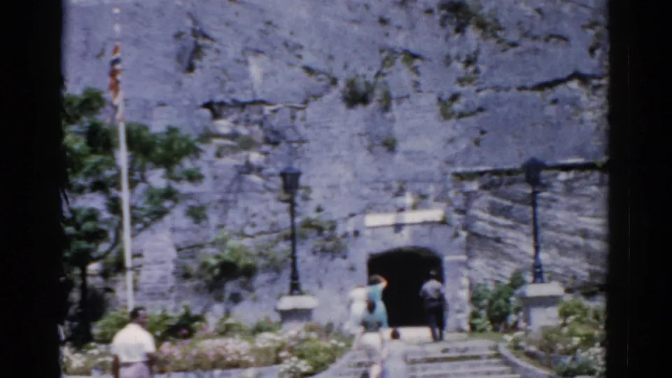 1975 Florida home-movie frame of visitors approaching an arched stone fortress entrance with steps, lamps, and flagpole nearby.