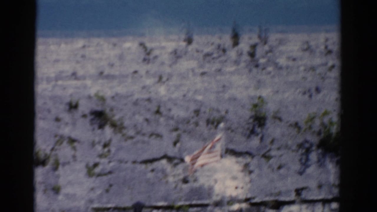 Small U.S. flag on pale rocky hillside with sparse shrubs and blue sky.