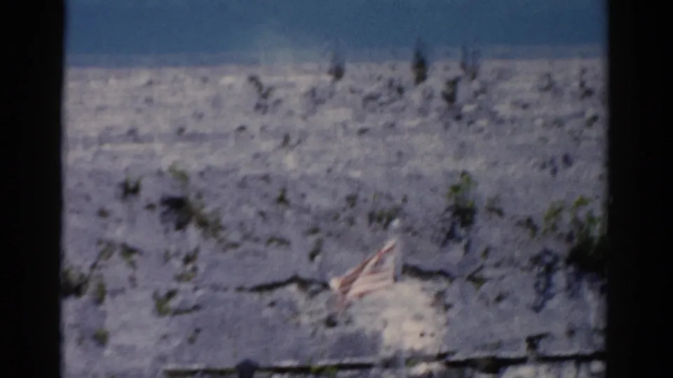 1975 Florida 8mm frame showing a small U.S. flag on a pale rocky hillside with scattered shrubs under blue sky.