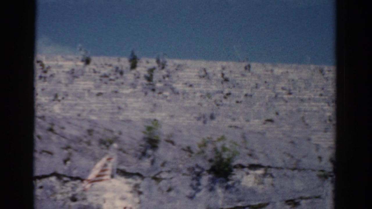 1975 Florida 8mm view of a small flag near cracked pale rock, low shrubs, and a clear blue sky.