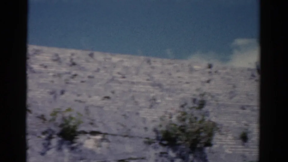1975 Florida 8mm frame of a pale rocky slope with shrubs, blue sky, and a small cloud; the flag is not prominent.
