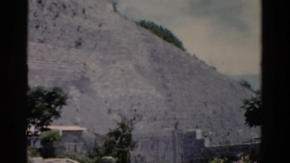 1975 8mm travel frame showing the sloped edge of a stepped stone structure with greenery and a small light-roofed building below.