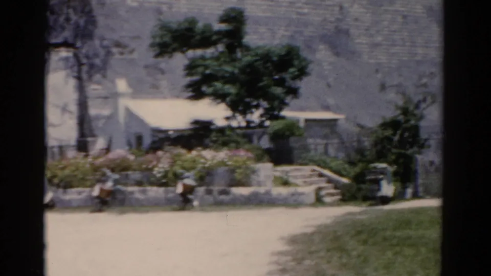 1975 8mm frame of a small white building, garden beds, trees, and a paved foreground below the stepped stone structure.