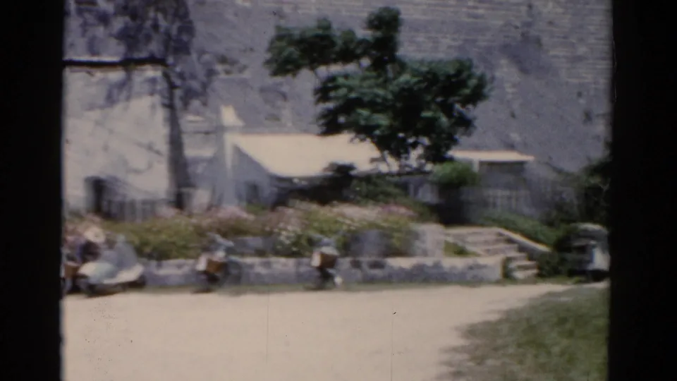 1975 Florida home-movie frame showing a sunlit street, low planter, weathered wall, tree, and people sitting near the curb.