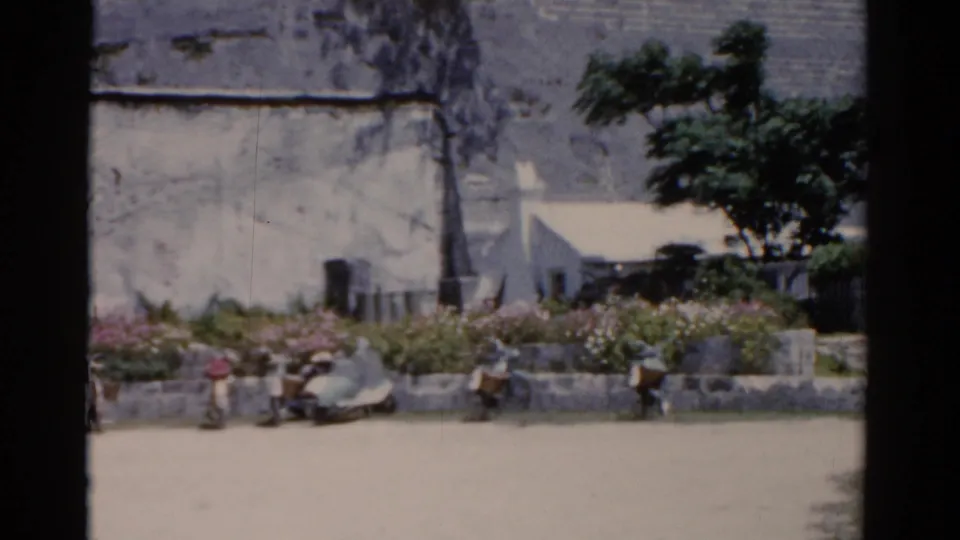 1975 Florida street scene with seated people, a white rounded car, flower beds, a tree, and a pale weathered wall.