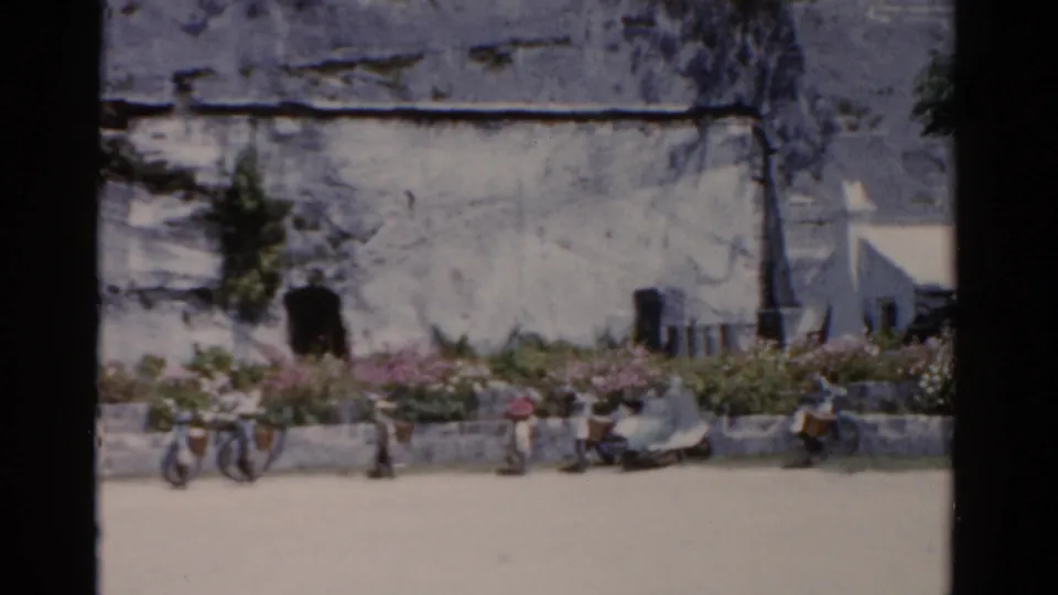 1975 Florida home-movie view of bicycles, seated people, flowers, and a weathered facade along a sunlit street.
