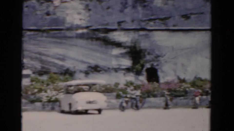 1975 Florida street frame with a white car in the foreground, bicycles at the curb, seated people, flowers, and a worn wall.