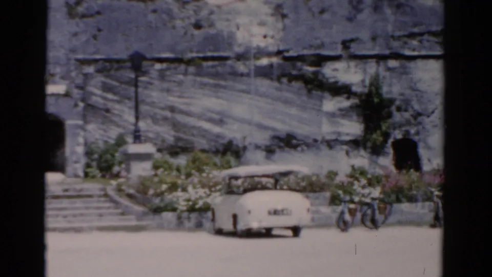 1975 Florida 8mm frame showing a white convertible beside stone steps, lampposts, planting beds, and a dark arched doorway.