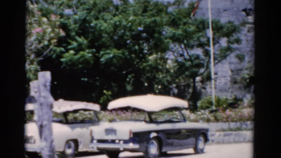 1975 Florida frame with two vintage cars parked near trees, planting beds, and the stone entrance area.