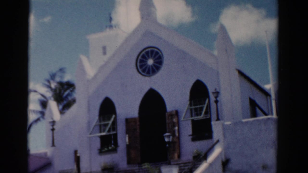 1975 Florida home-movie frame of a white church entrance with Gothic-style arches, circular rose window, open shutters, and bright sky.