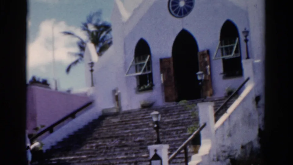 1975 Florida 8mm frame widening to show the church facade, broad stone staircase, lampposts, palm foliage, and a pink neighboring wall.