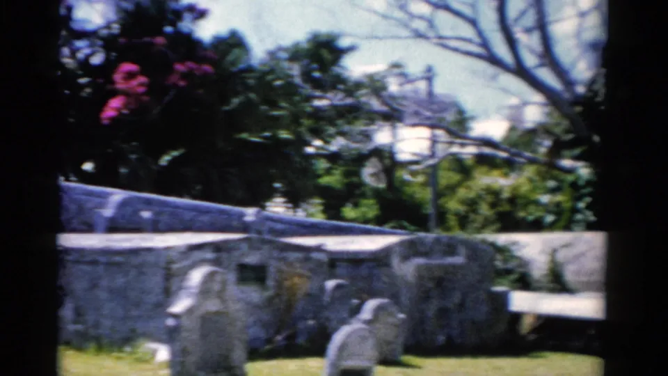 Florida 1975 8mm frame with stone cemetery structures, grave markers, grass, trees, and pink flowering foliage.