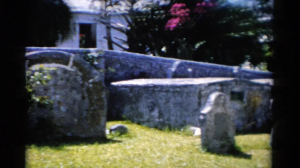 1975 Florida home-movie frame with a curving stone wall, gravestones, lawn, trees, and pink flowers in strong sun.