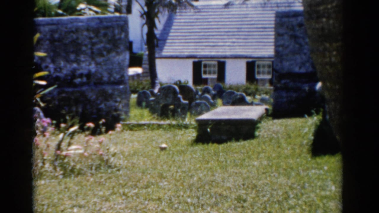 1975 Florida frame of a sunlit cemetery lawn with clustered headstones, palms, stonework, and a white building behind the graves.