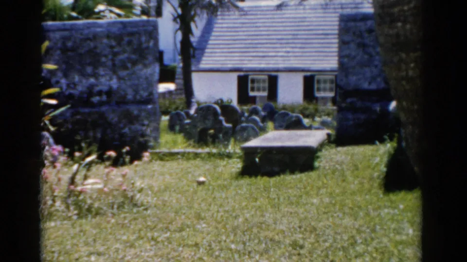 1975 Florida frame of a sunlit cemetery lawn with clustered headstones, palms, stonework, and a white building behind the graves.