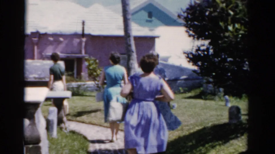 1975 Florida home-movie frame showing three women walking away through a tropical cemetery with gravestones, palms, and a small building ahead.
