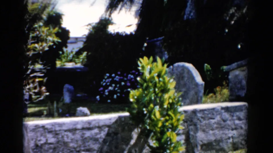 1975 Florida frame showing a shaded cemetery corner with stone wall, upright grave markers, shrubs, flowers, and dense tropical foliage.