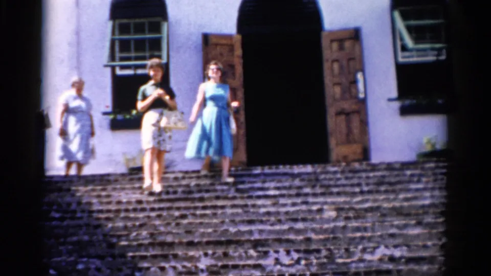 Three women in 1970s dresses and skirt walk down stone steps outside a pale building in Florida.